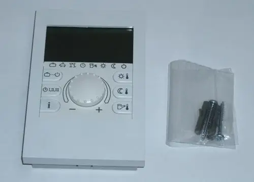 Weißes Bedienelement mit Display und verschiedenen Symbolen, darunter Einstellknopf und Funktionstasten, neben einer Tüte mit Montagematerial von Daikin.